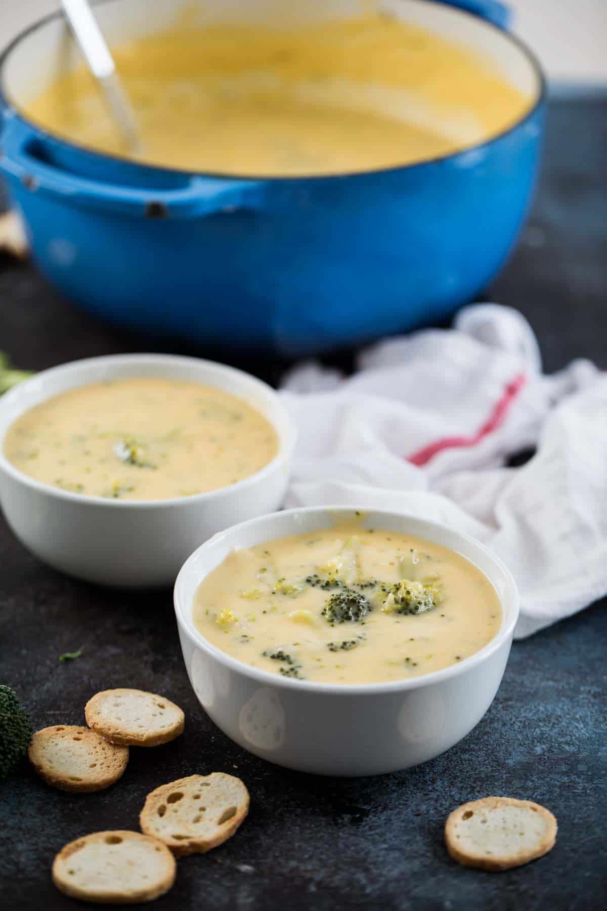 two bowls of broccoli cheese soup with a pot in the back