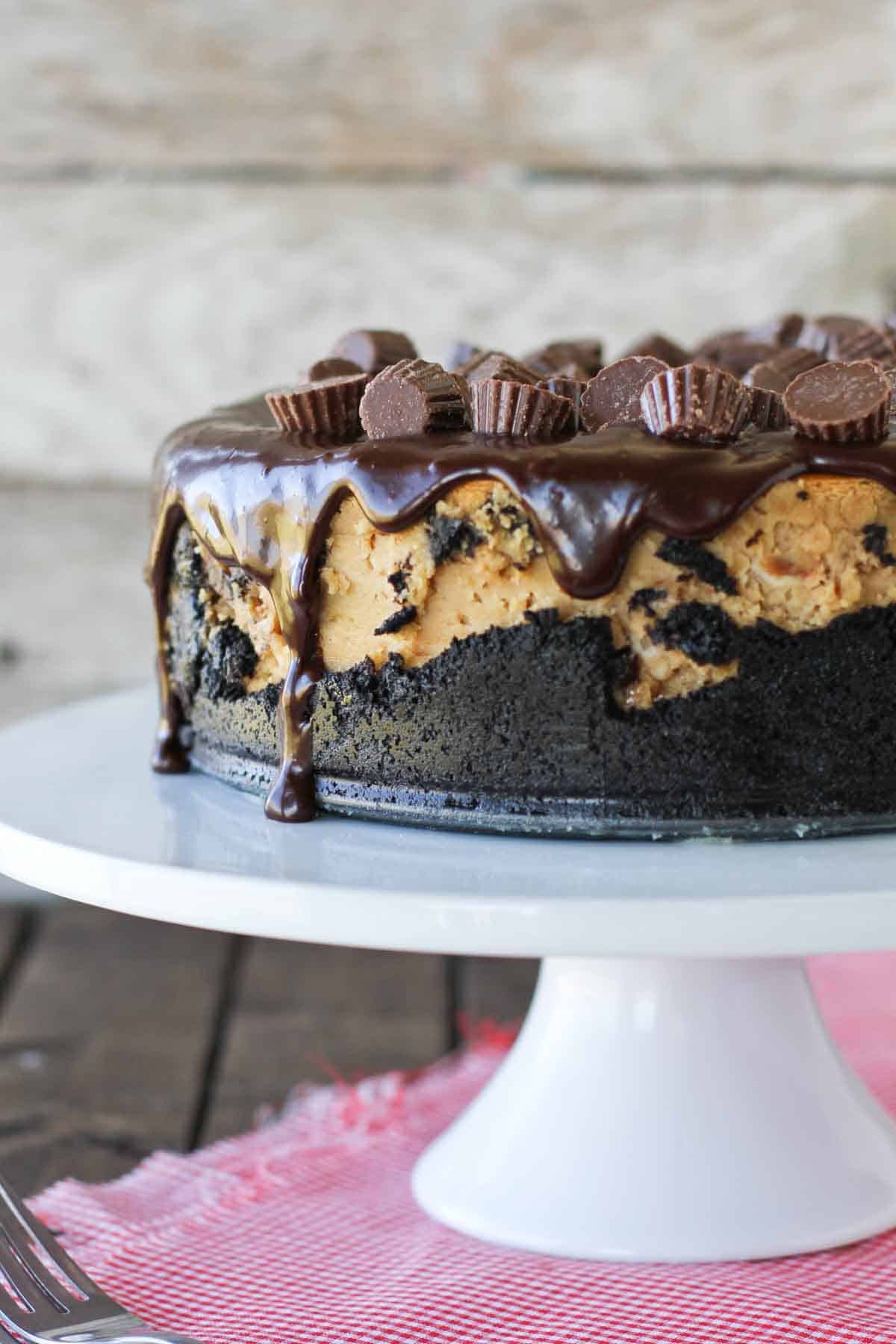 Rich and decadent, this Reese’s Peanut Butter Cheesecake Recipe starts with a cookie crust, is filled with Reese’s Peanut Butter Cups, then topped with a chocolate ganache and even more peanut butter cups. full Reese's Peanut Butter Cheesecake on a cake stand.