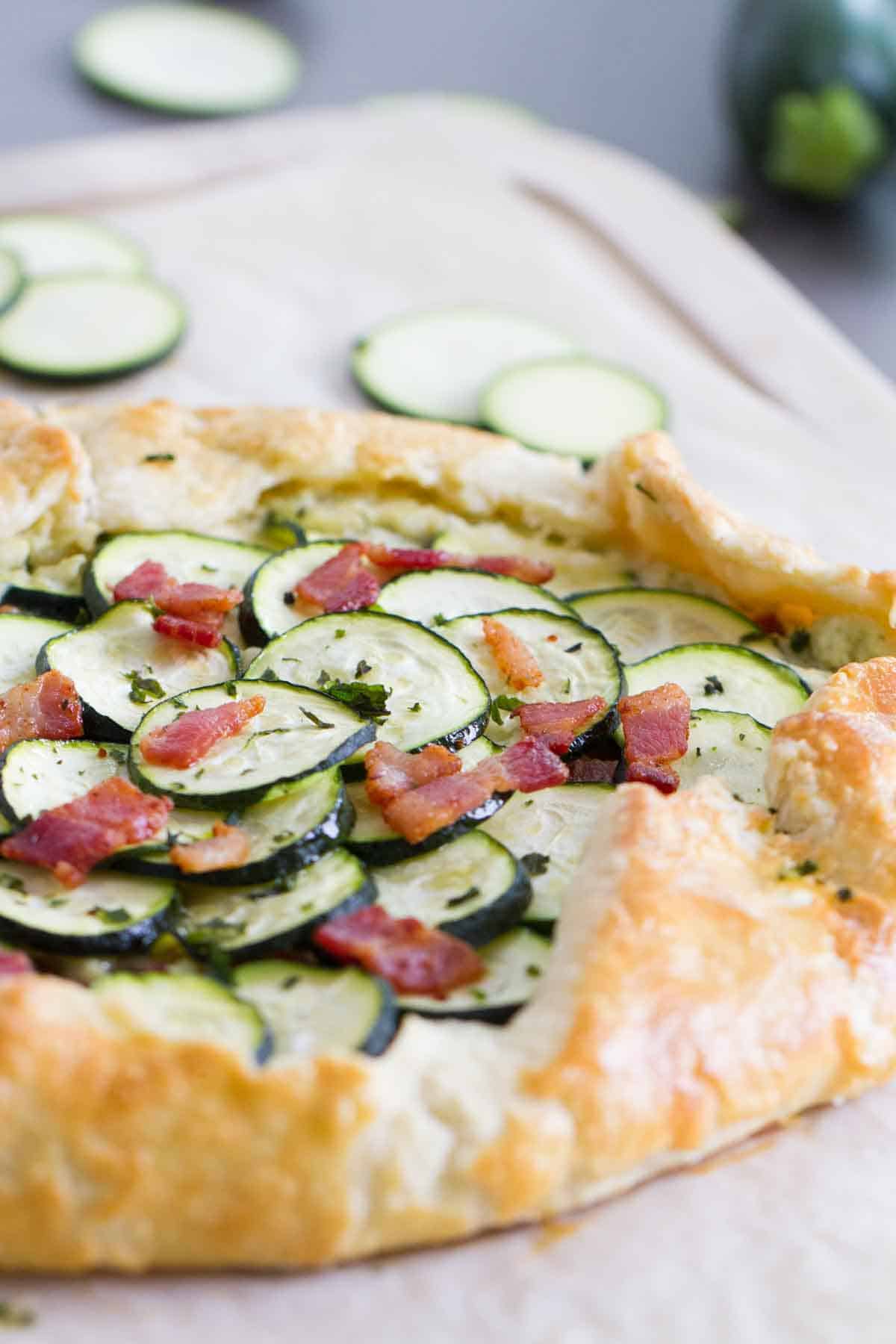 A tender, flaky crust is filled with a creamy ricotta filling, then topped with bacon and zucchini in this easy Zucchini Tart with Ricotta and Bacon that is perfect for a luncheon or a light dinner. Full zucchini tart with ricotta and bacon on a cutting board