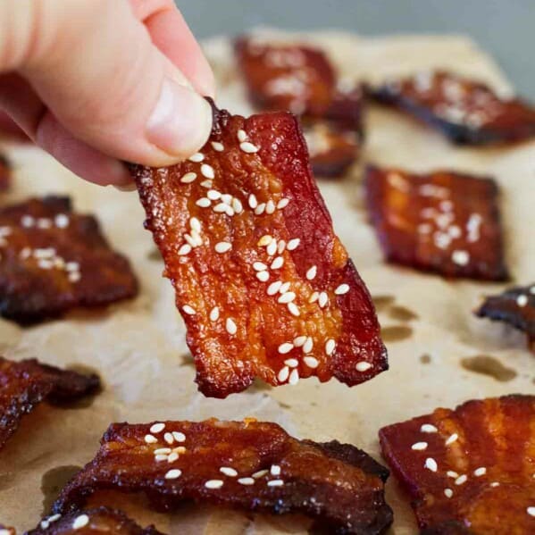 Fingers holding a piece of bacon seasoned with brown sugar and sriracha.