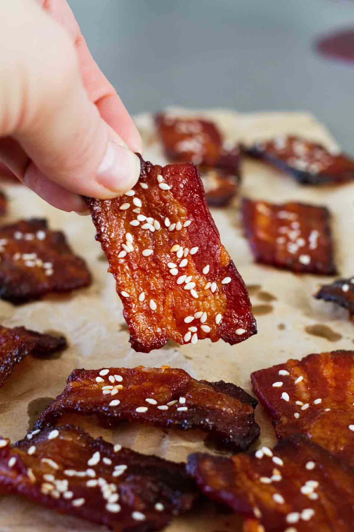 Fingers holding a piece of bacon seasoned with brown sugar and sriracha.