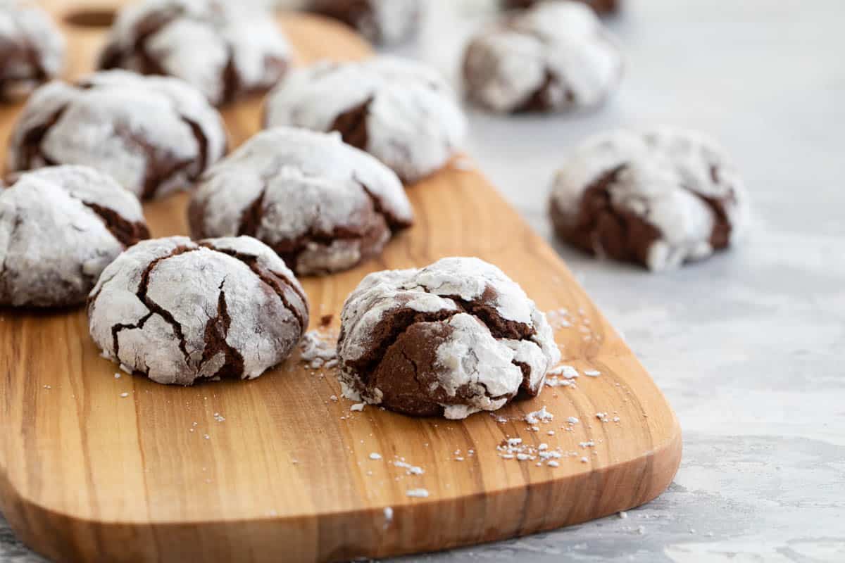 How to make Chocolate Crinkle Cookies - fudgy, brownie-like cookies that are coated in powdered sugar. These are a classic for a reason! Puffy chocolate crinkle cookies with powdered sugar placed on a cutting board.