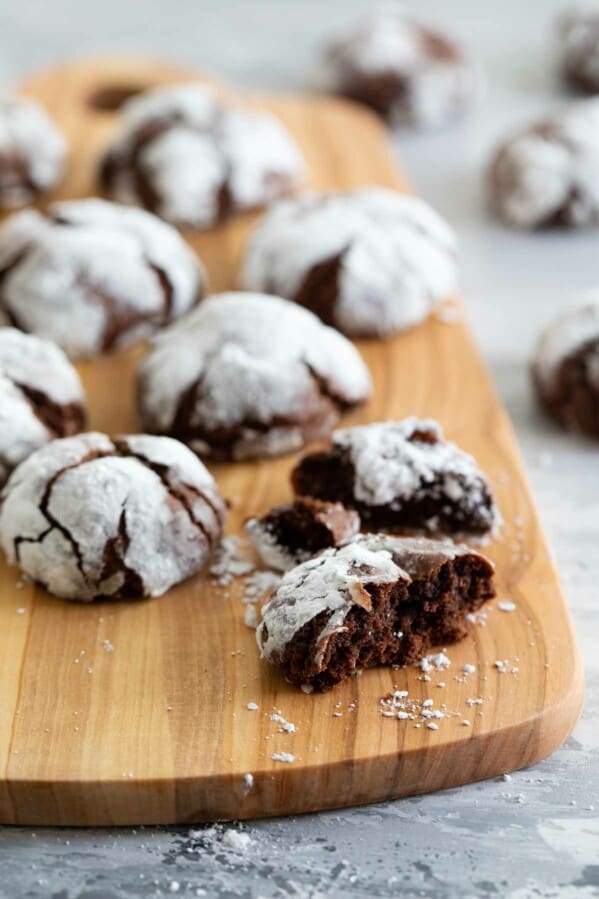 cutting board with chocolate crinkle cookies, with one cookie broken into pieces.