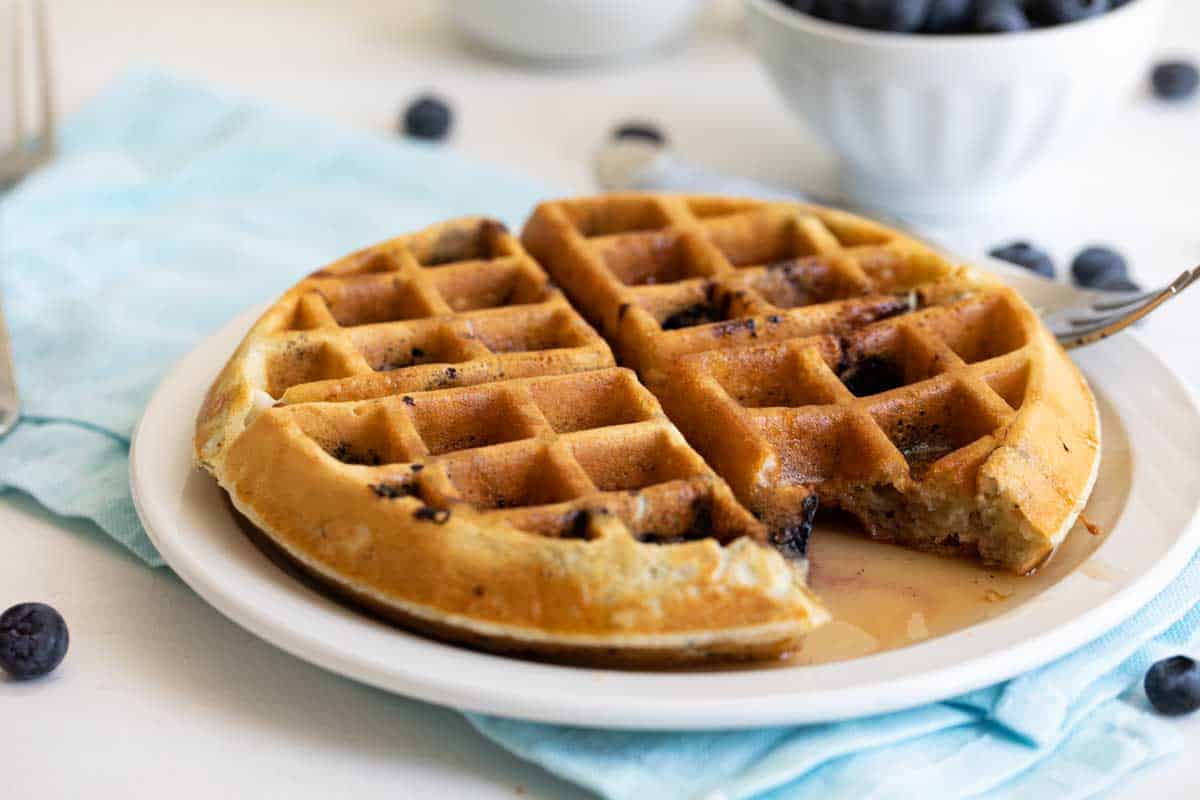 blueberry waffle on a white plate with a fork and a bite taken from the waffle
