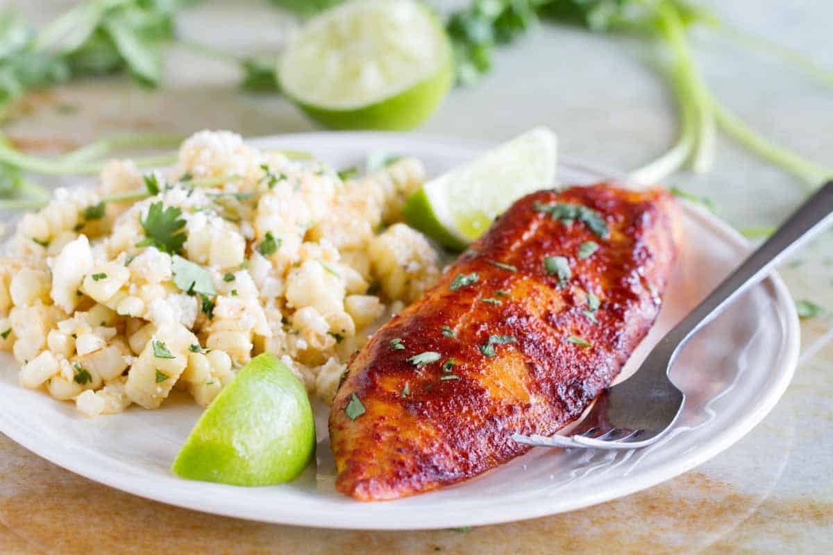 White plate with taco seasoned grilled fish, corn salad and lime wedges