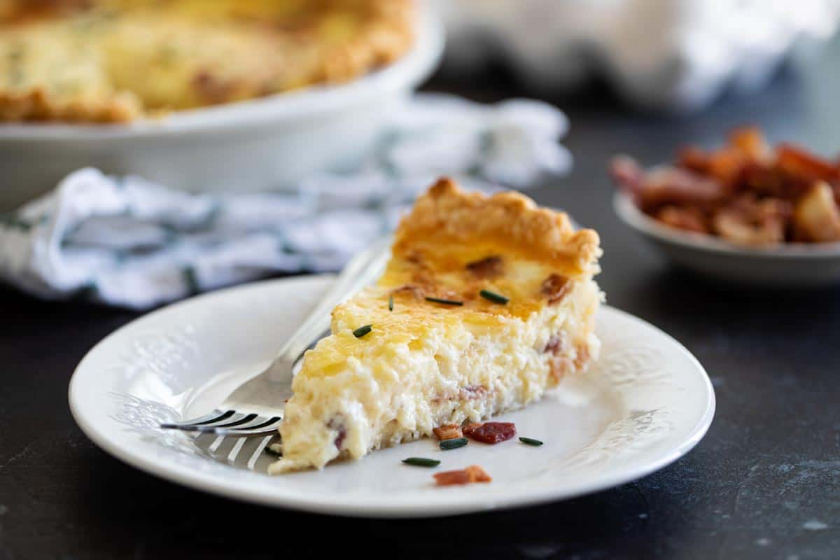 Perfect for breakfast, brunch or dinner, this classic Quiche Lorraine recipe is filled with bacon and cheese. Add a flaky pie crust and you have a family favorite. slice of quiche lorraine with chopped chives on a plate