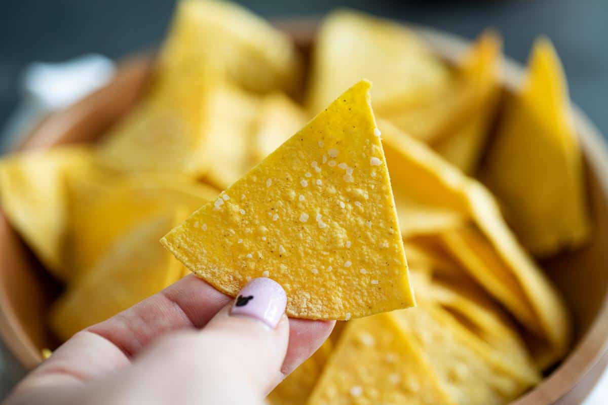 Crispy and salty, these Homemade Tortilla Chips are so easy to make and so much better for you than store bought. They are the perfect afternoon snack. single tortilla chip with bowl of tortilla chips behind