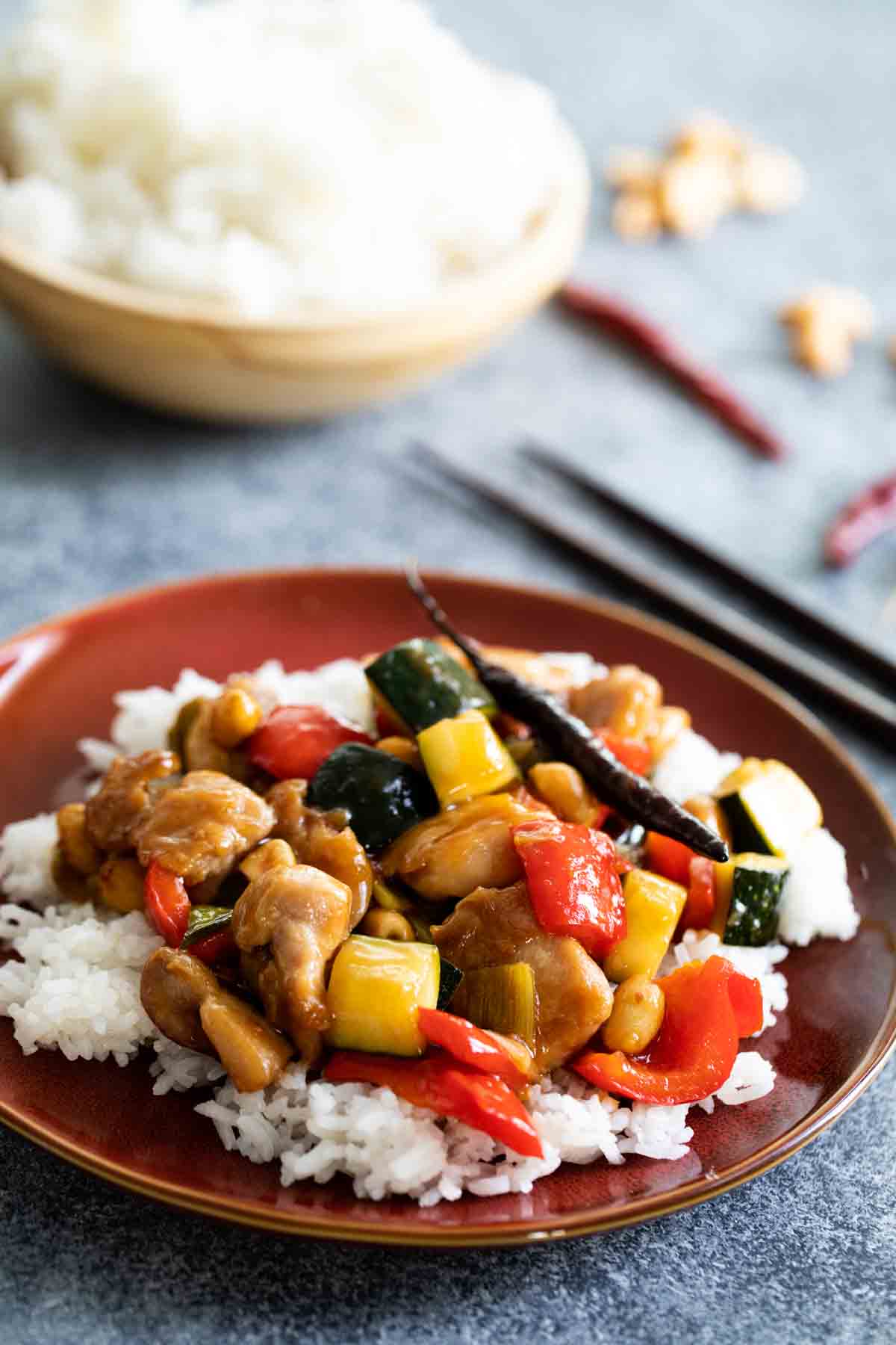 plate of kung pao chicken over rice with rice and chopsticks in the background.