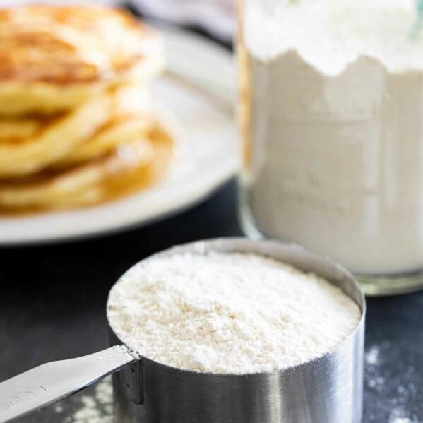 pancake mix in a measuring cup with pancakes and more mix behind