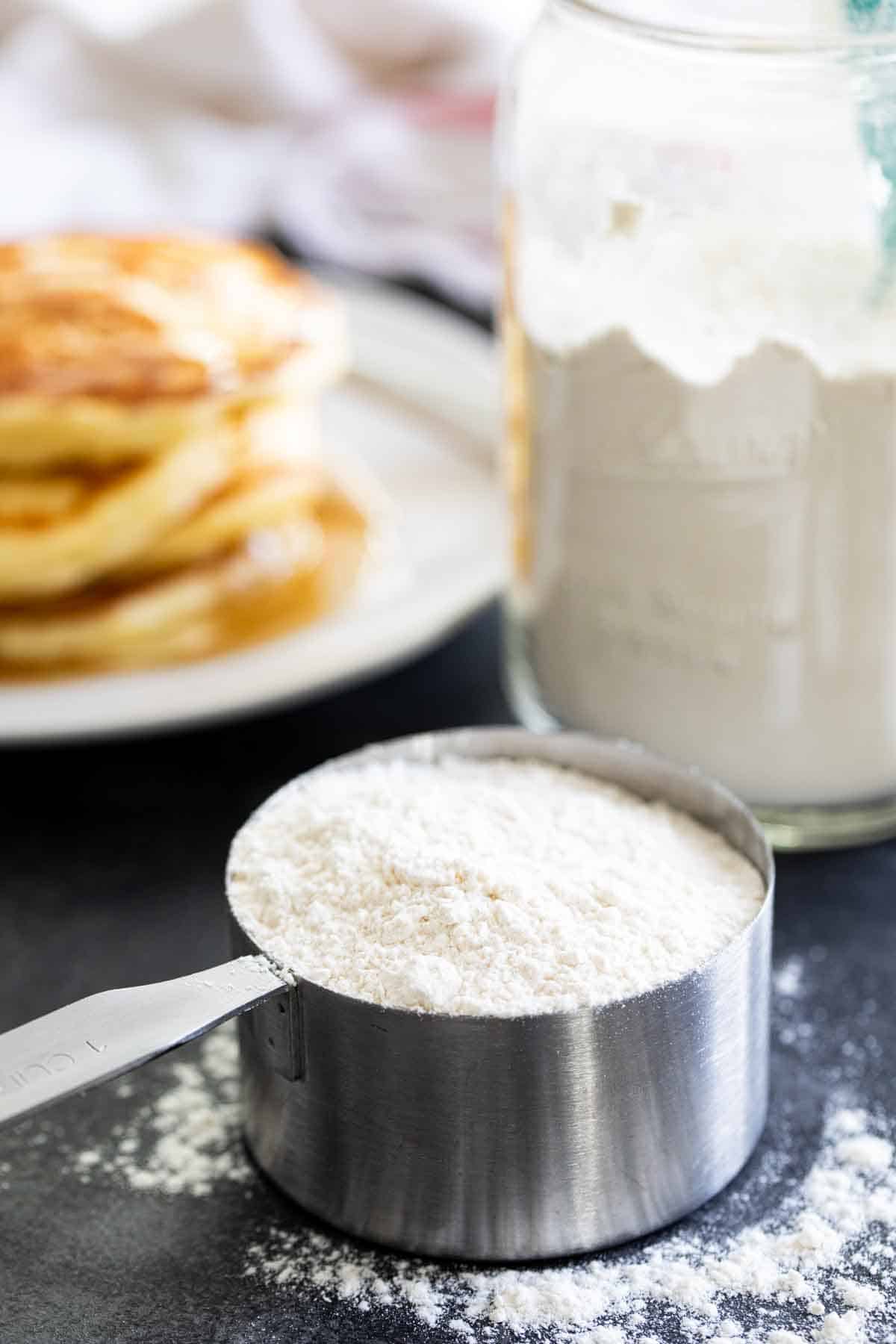 pancake mix in a measuring cup with pancakes and more mix behind