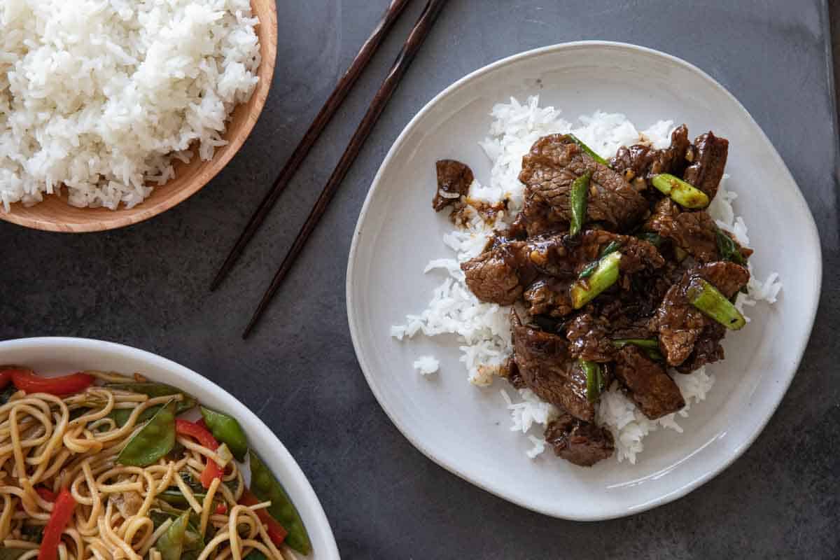 overhead view of mongolian beef over rice on a plate with rice and lo mein noodles on the side