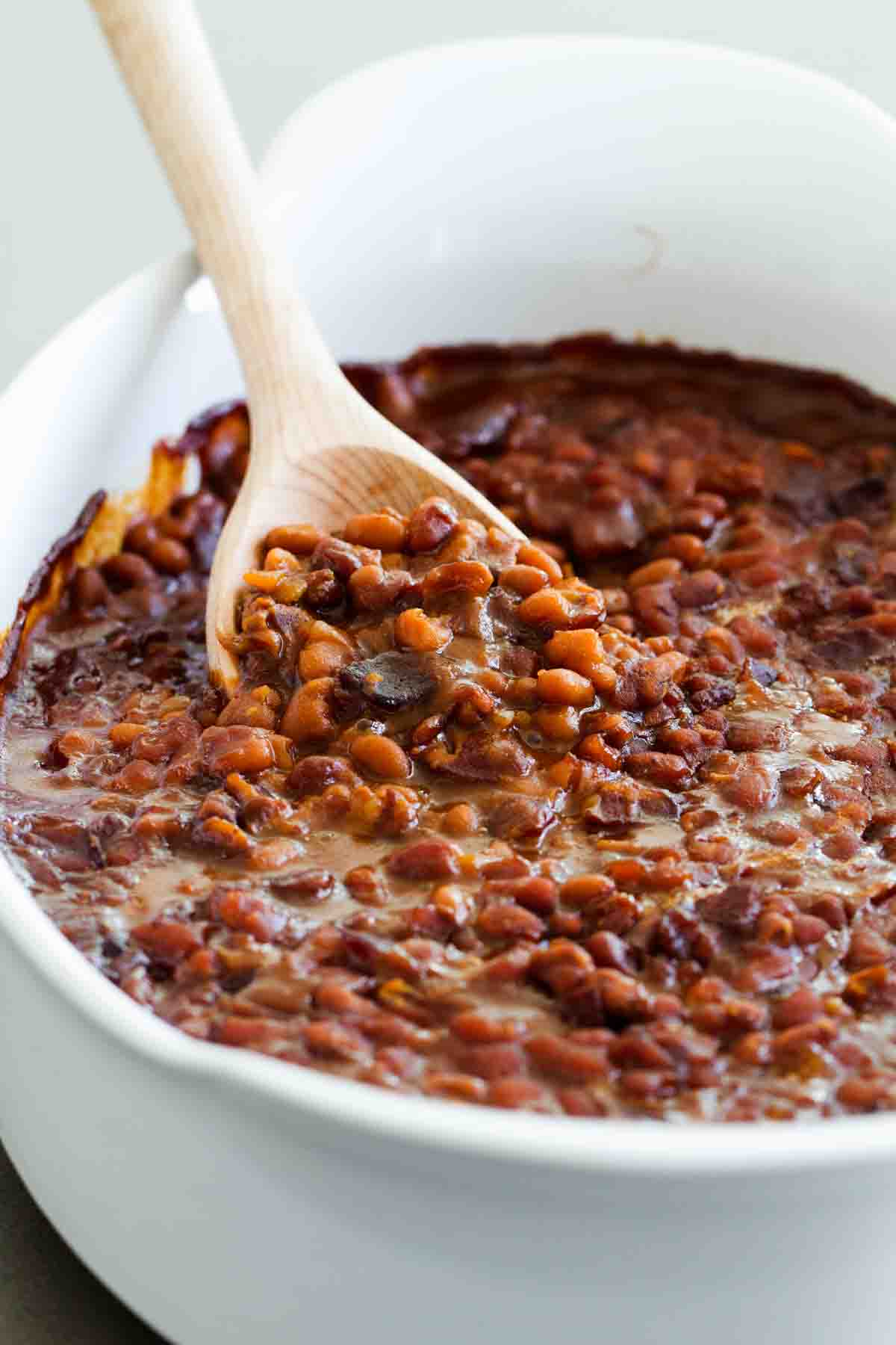 Baked beans with bacon in a casserole dish with a wooden spoon to scoop