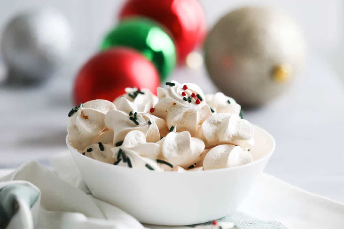 Crunchy, light and sweet, these classic Meringue Cookies only take 5 ingredients and are perfect for your holiday cookie plates. bowl filled with meringue cookies with Christmas ornaments in the background