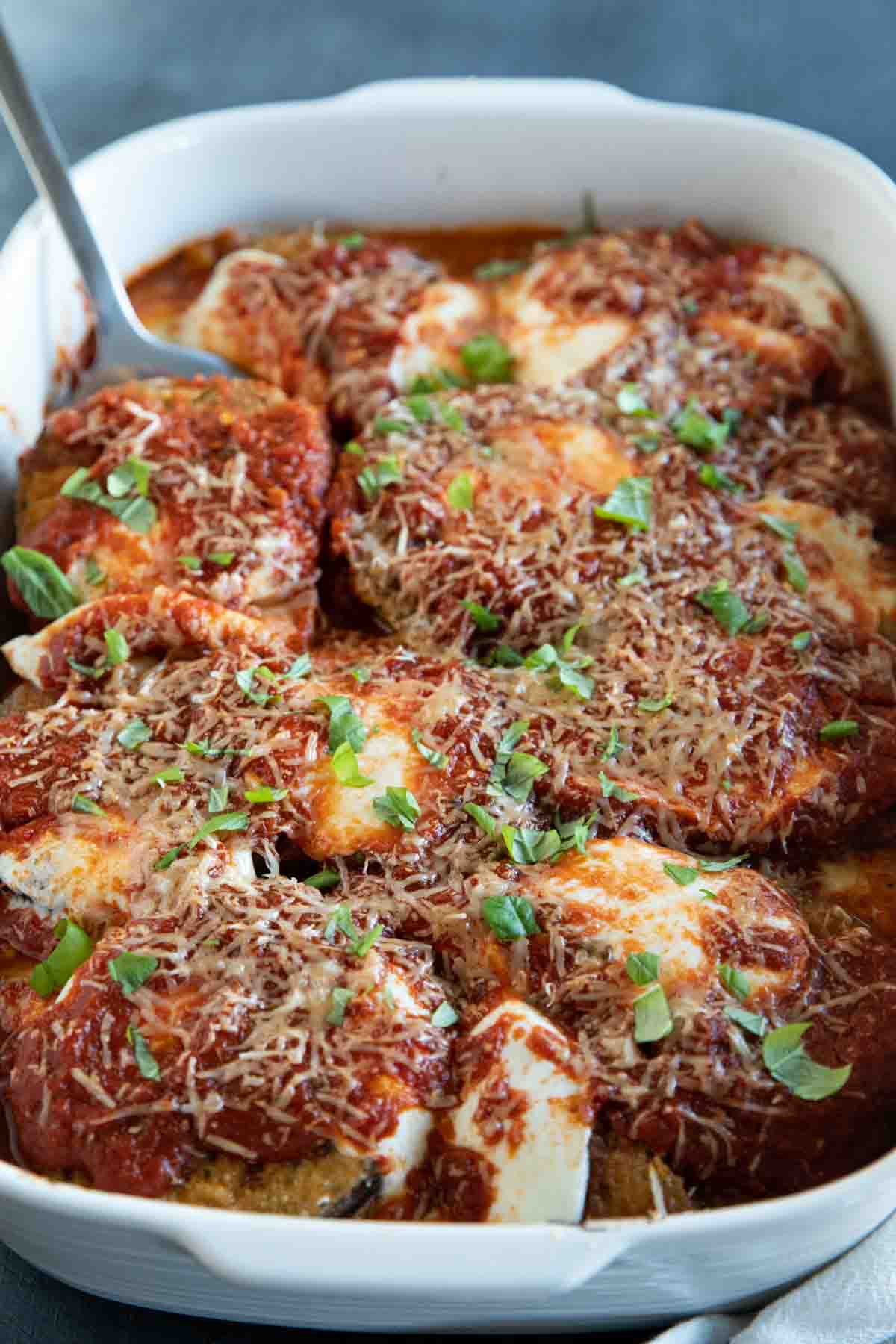 white casserole dish with eggplant parmesan with spatula.