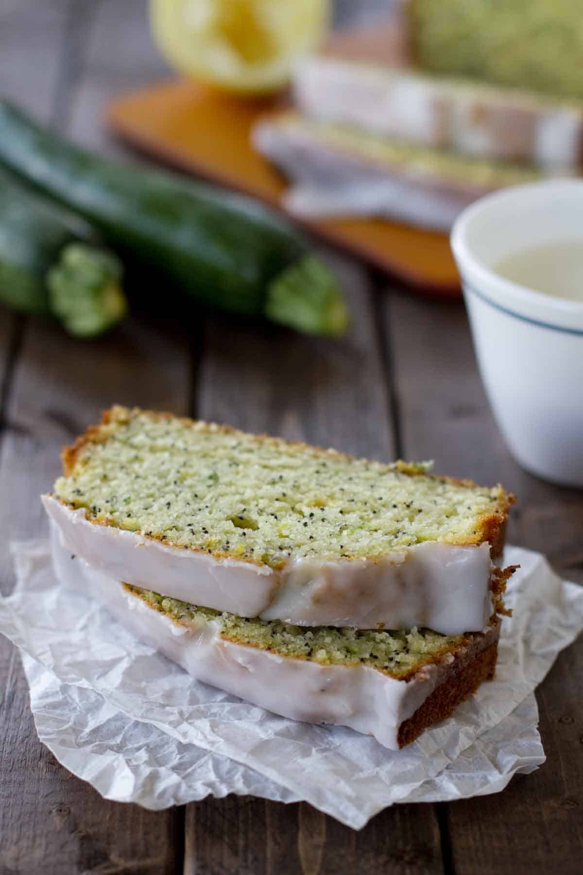 Two slices of lemon poppy seed zucchini bread on paper with zucchini in the background.