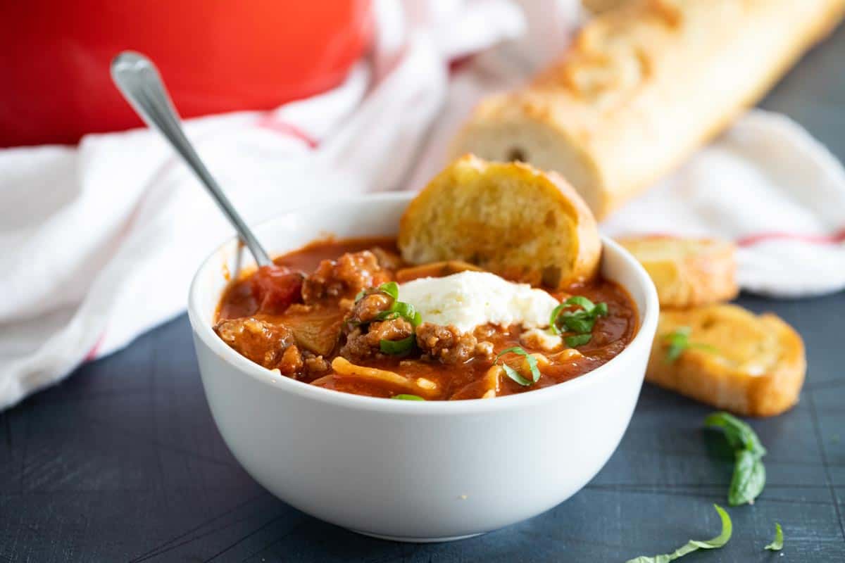 white bowl with lasagna soup topped with ricotta cheese with a spoon and bread