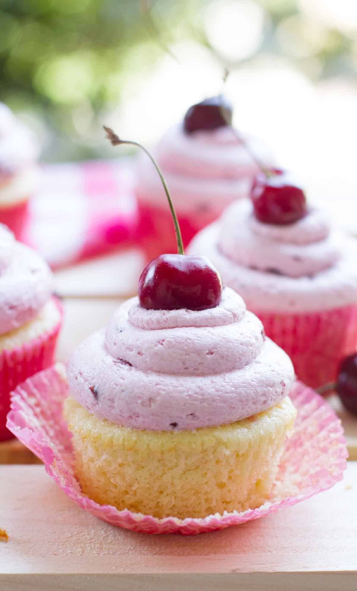 almond cupcake with the paper pulled back showing texture, topped with fresh cherry frosting.