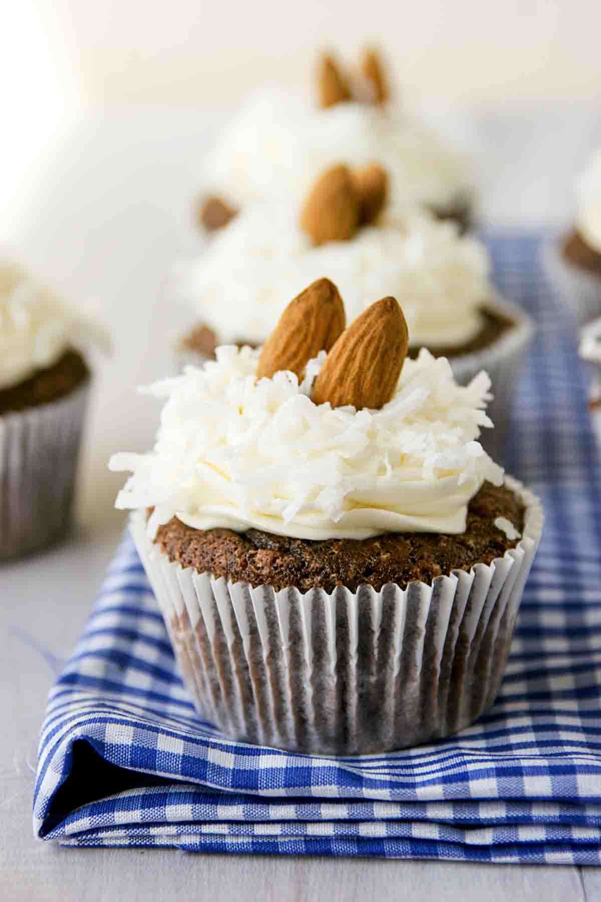 Love coconut and chocolate? These Almond Joy Cupcakes have a chocolate and coconut cupcake that is topped with a coconut buttercream, more coconut, and almonds. Almond Joy Cupcakes in a row on top of blue and white fabric