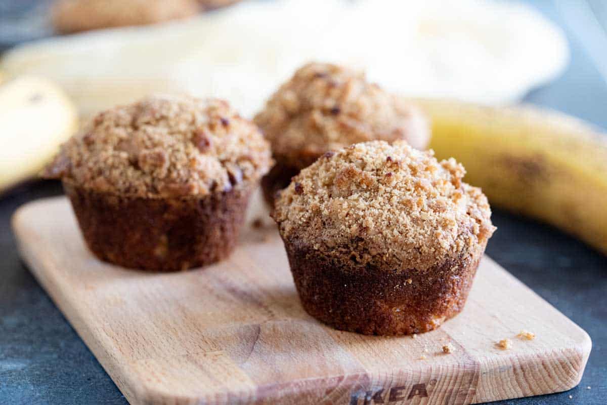 Three banana muffins with crumb topping on a cutting board.