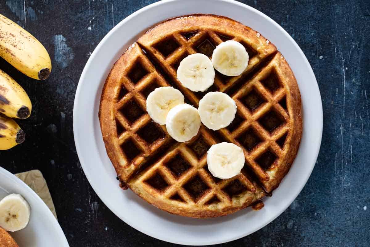 Turn those over-ripe bananas into breakfast with these easy Banana Waffles. For extra banana flavor, serve with more bananas on top! Banana waffle on a plate with fresh bananas on top.