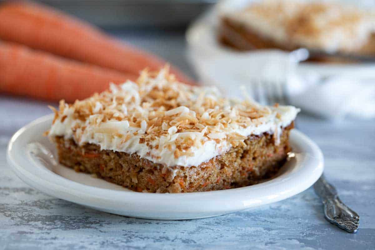 slice of carrot sheet cake with toasted coconut on a plate with carrots in the background