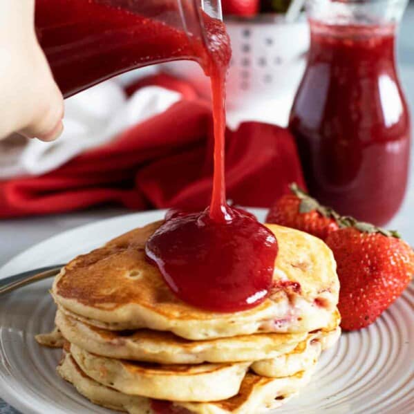 pouring strawberry syrup onto a stack of pancakes