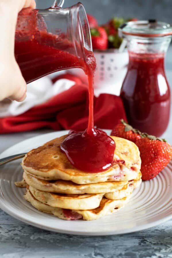 pouring strawberry syrup onto a stack of pancakes