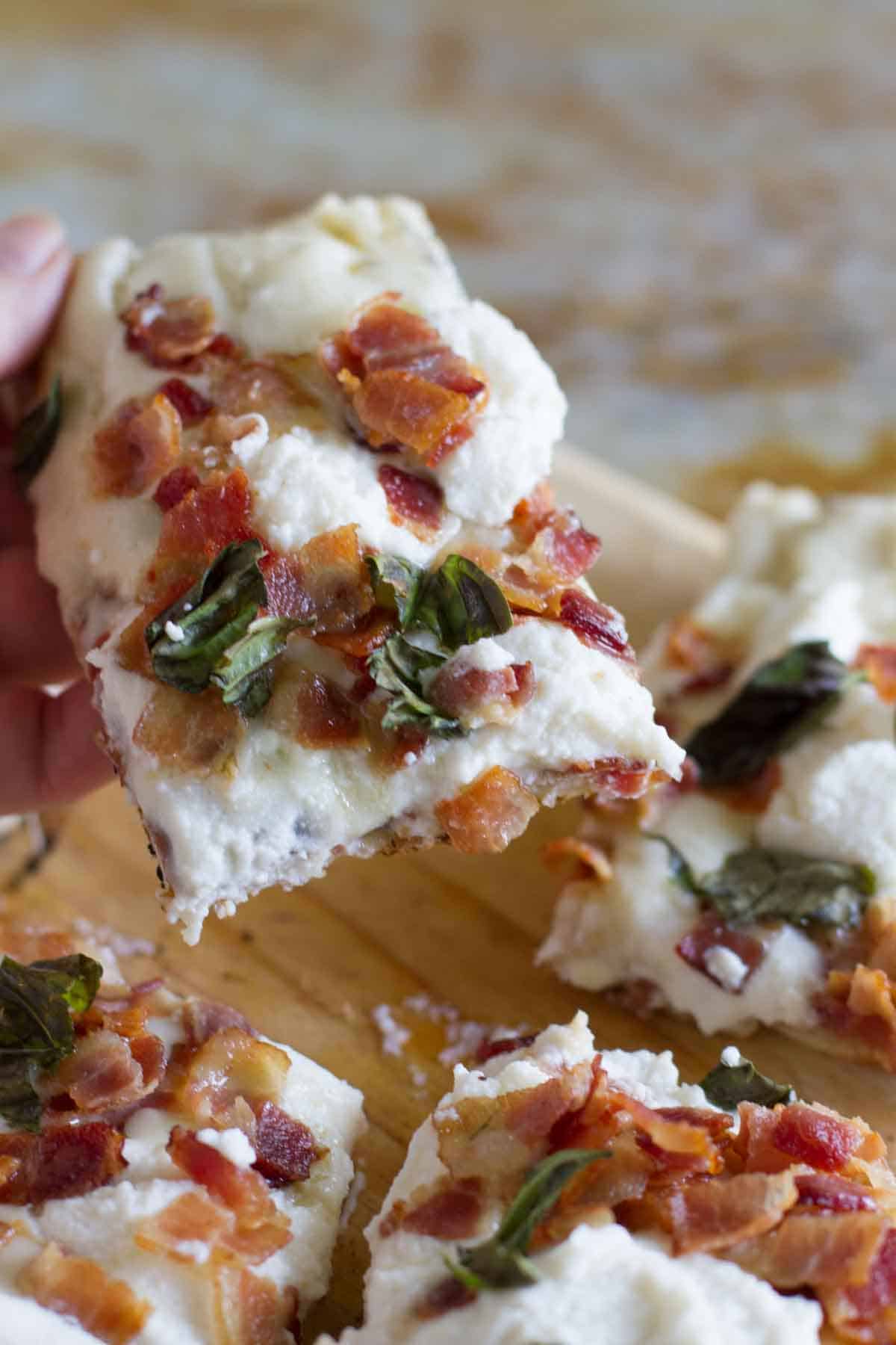 holding a slice of grilled white pizza with bacon and basil