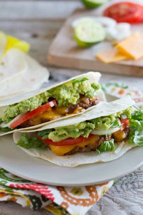 two Cheeseburger Tacos on a plate with toppings in the background