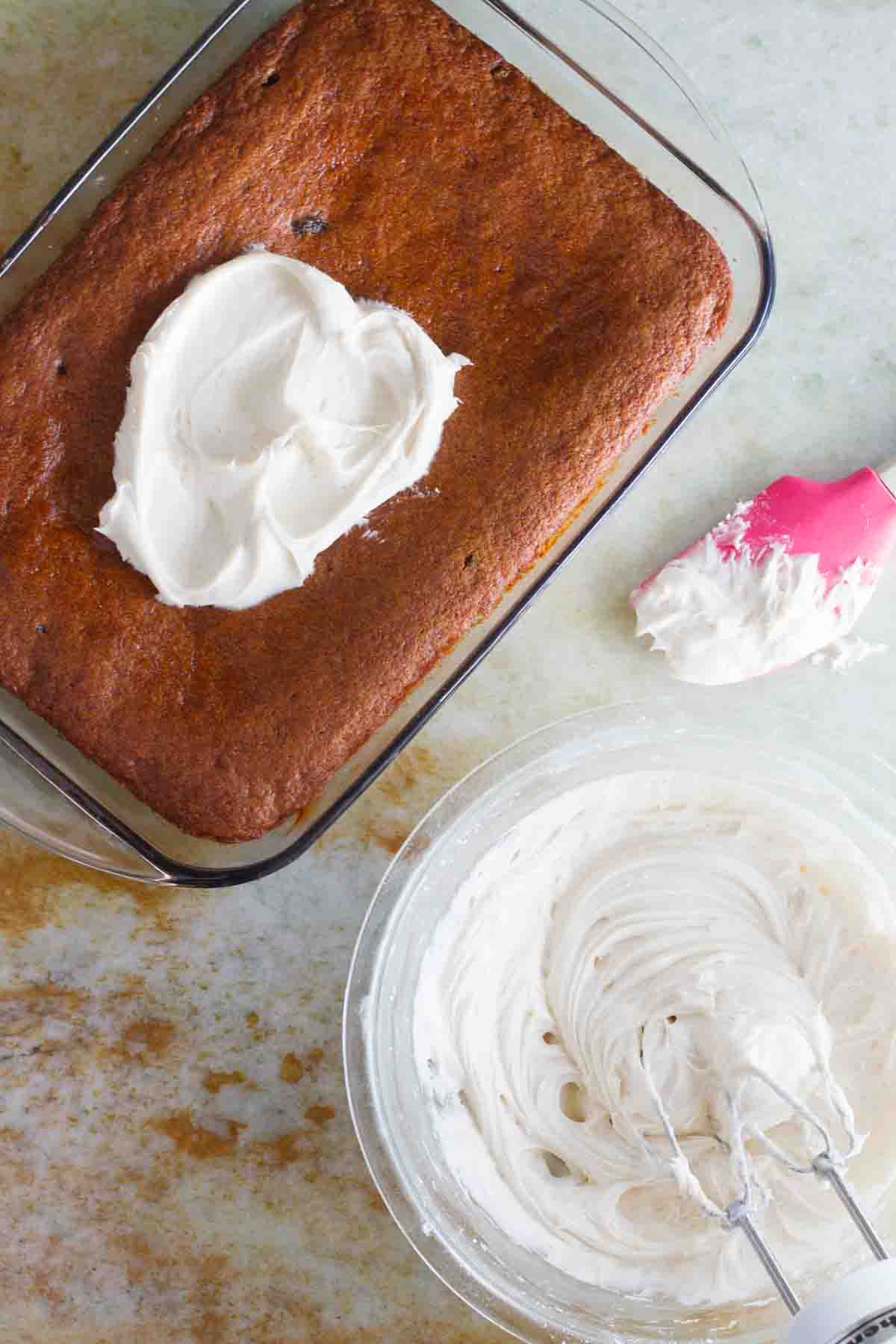 Bowl with cream cheese icing and pan with cake that has a dollop of icing on top