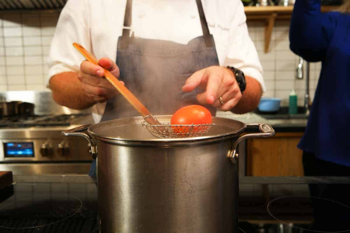 Man getting a tomato from a large pot with a straining spoon.