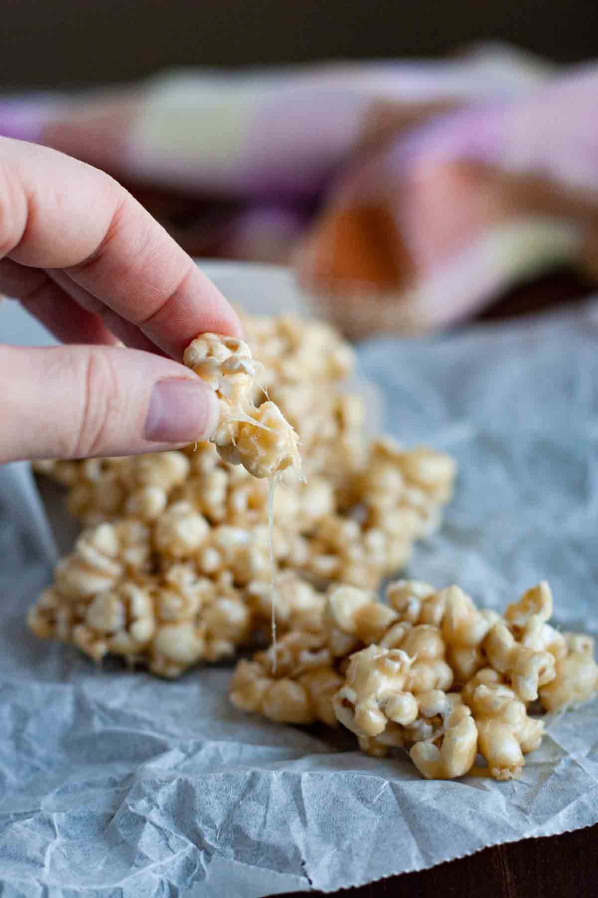Ooey and gooey, this Marshmallow Caramel Popcorn is coated with a marshmallow and caramel mixture, making it addictive and delicious. fingers holding a piece of Marshmallow Caramel Popcorn