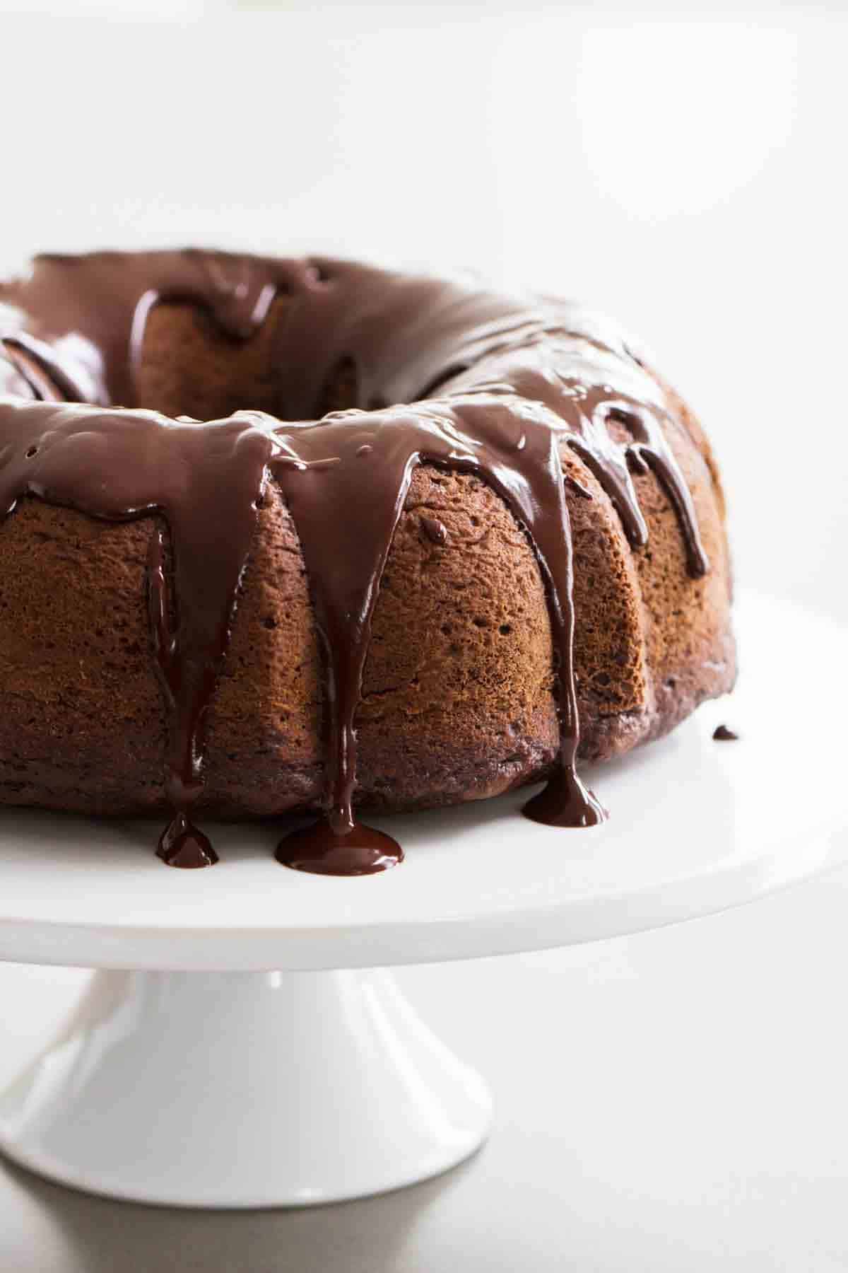 full chocolate bundt cake with chocolate icing on a white cake stand.