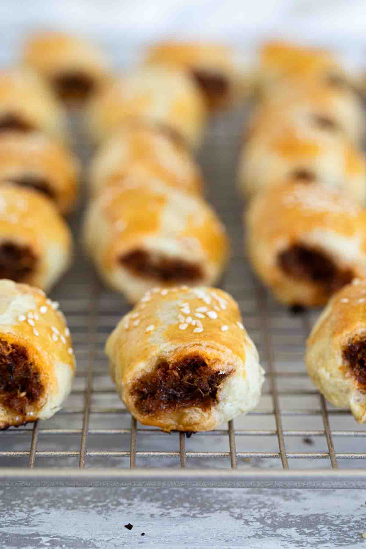 A popular British snack, these simple Sausage Rolls have puff pastry that is filled with a sausage mixture and baked. Sausage Rolls on a cooling rack.