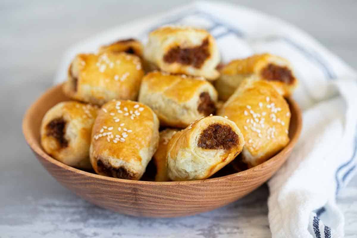 A popular British snack, these simple Sausage Rolls have puff pastry that is filled with a sausage mixture and baked. Sausage Rolls in a wooden bowl.