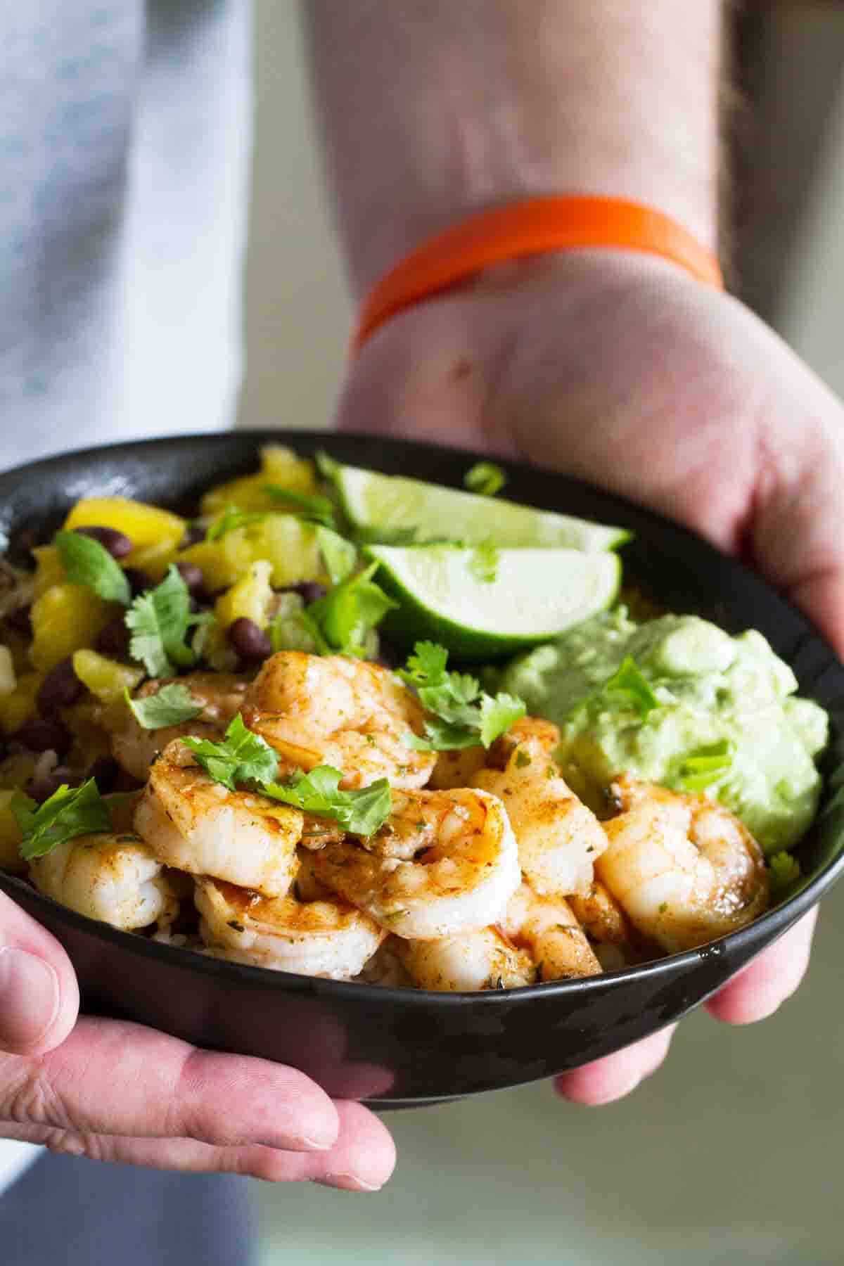 Hands holding a bowl filled with shrimp, rice, pineapple, mango, and avocado.