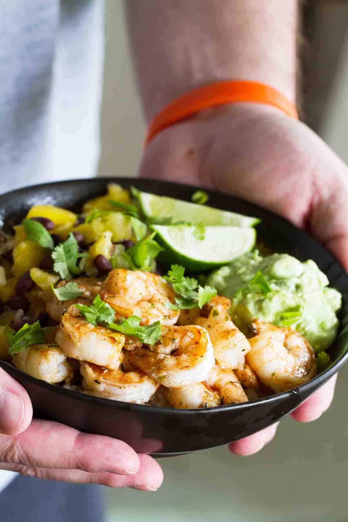 Sweet, spicy and savory – these Spicy Shrimp Bowls have it all going on! Made with coconut rice, beans, pineapple, mango, avocado and spicy shrimp, these bowls are begging to be made over and over again! Hands holding a bowl filled with shrimp, rice, pineapple, mango, and avocado.