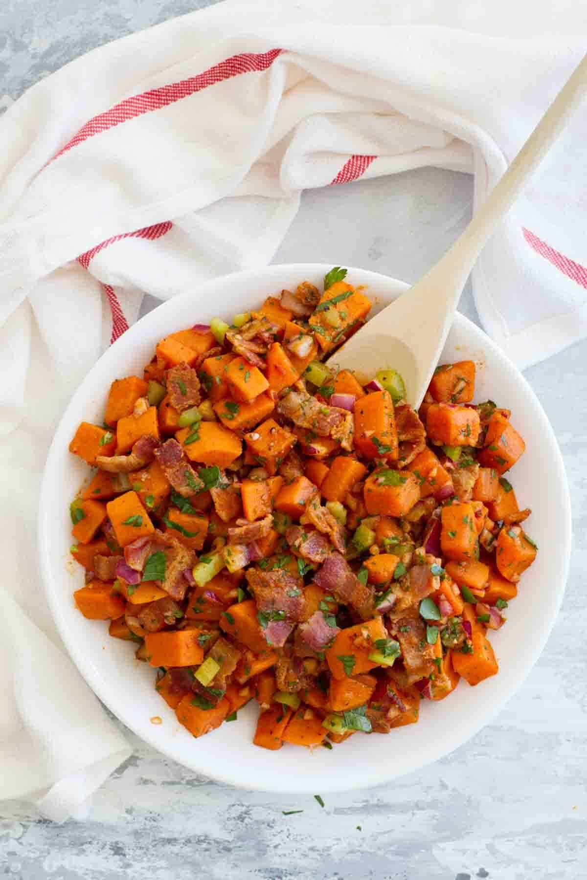 Sweet Potato Salad with Bacon in a white bowl with a wooden spoon.