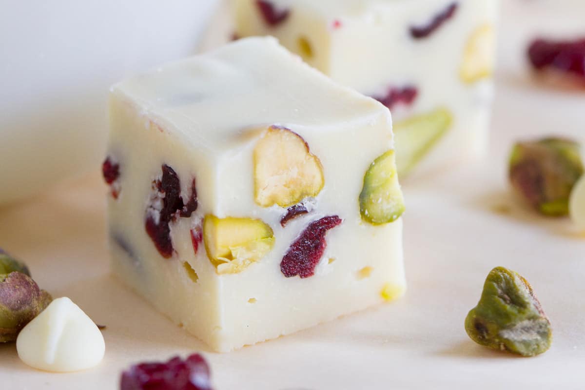 Square of creamy cranberry pistachio fudge surrounded by nuts, cranberries, and a white chocolate chip.