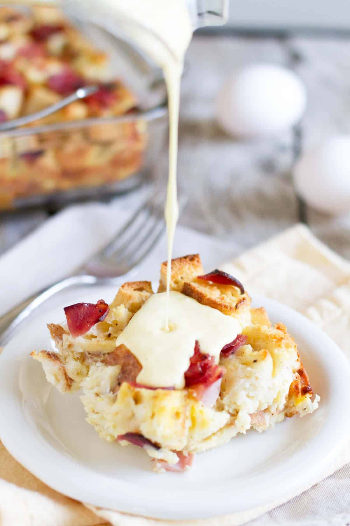 Pouring hollandaise sauce over the top of a piece of eggs benedict casserole.