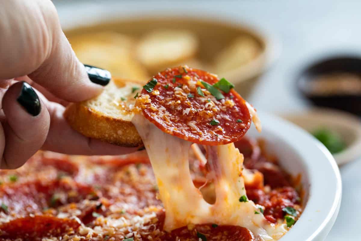 Cheese pull of pepperoni pizza dip on a baguette slice.