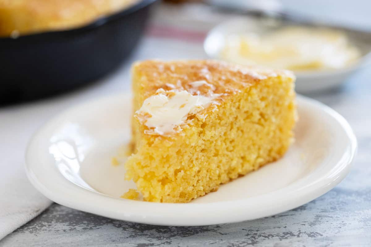 As easy as a box mix but a lot tastier, this homemade Cornbread Recipe is the perfect side dish. Serve it up with chili, beans, or with a pat of butter and a drizzle of honey. Slice of cornbread topped with butter on a plate.