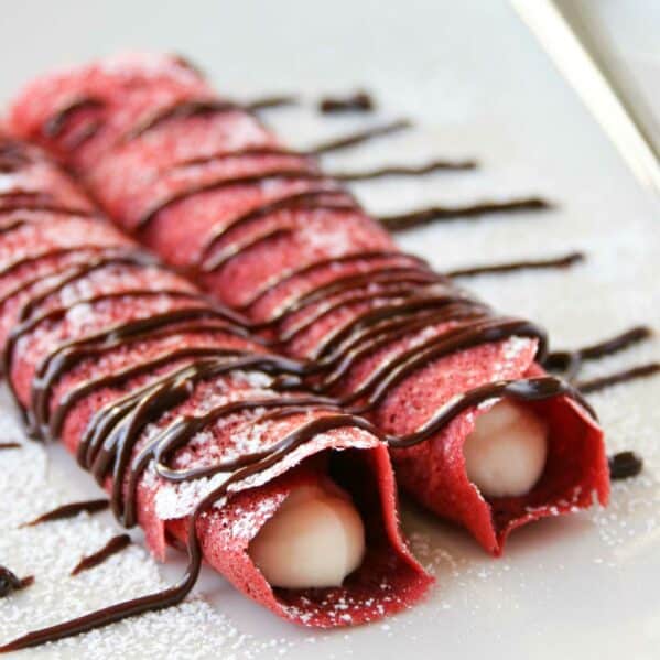 Two rolled red velvet crepes filled with cream cheese filling and drizzled with chocolate.