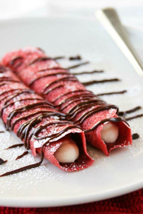 Two rolled red velvet crepes filled with cream cheese filling and drizzled with chocolate.