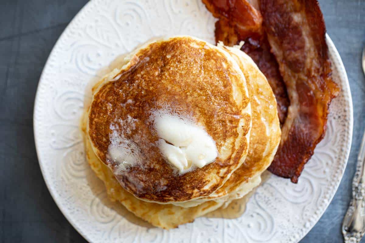 Stack of buttermilk pancakes topped with butter and syrup with bacon on the side.