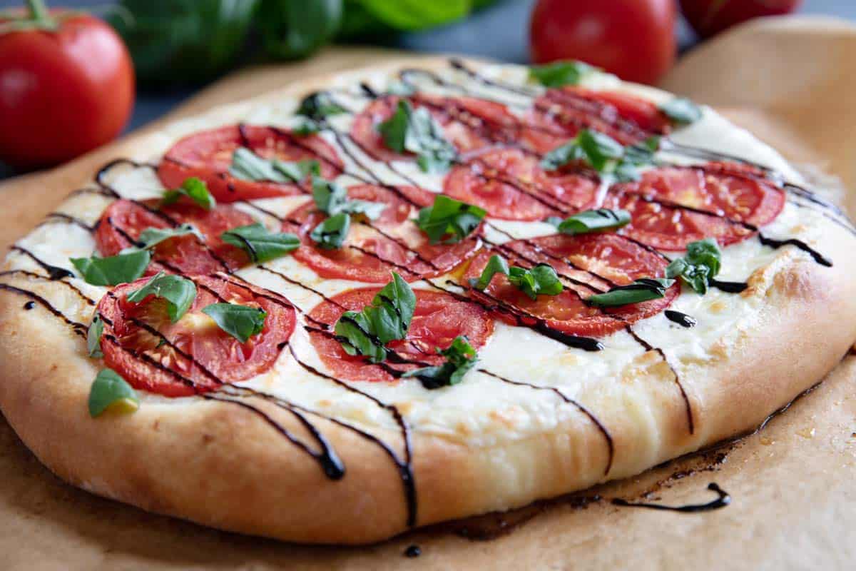 Caprese Pizza topped with fresh tomatoes and balsamic glaze with extra ingredients in the background.