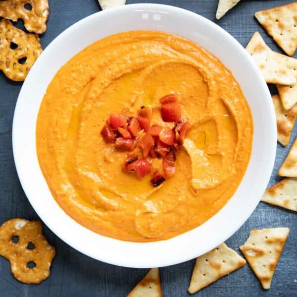 Bowl of roasted red pepper hummus surrounded by pitas, pita chips, and pretzels.
