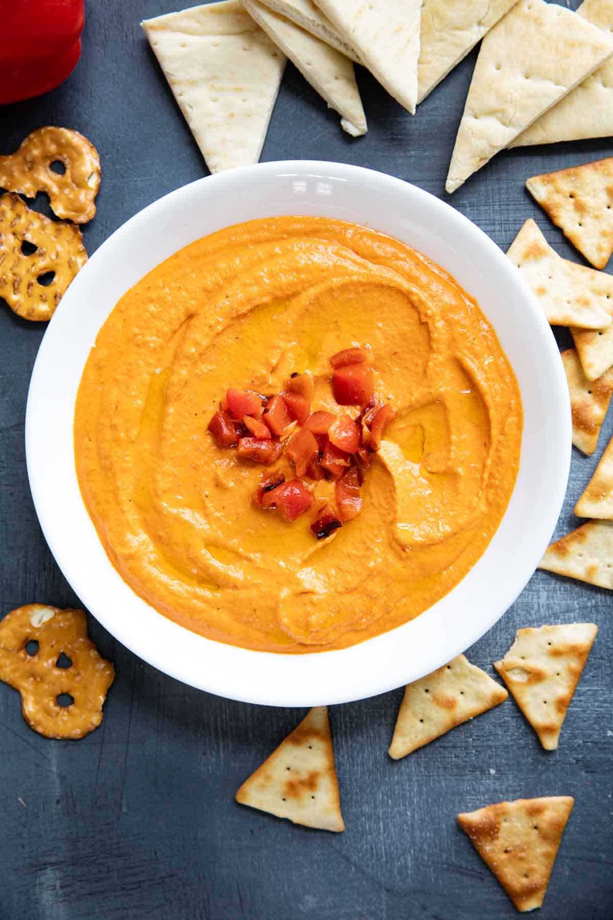 Bowl of roasted red pepper hummus surrounded by pitas, pita chips, and pretzels.