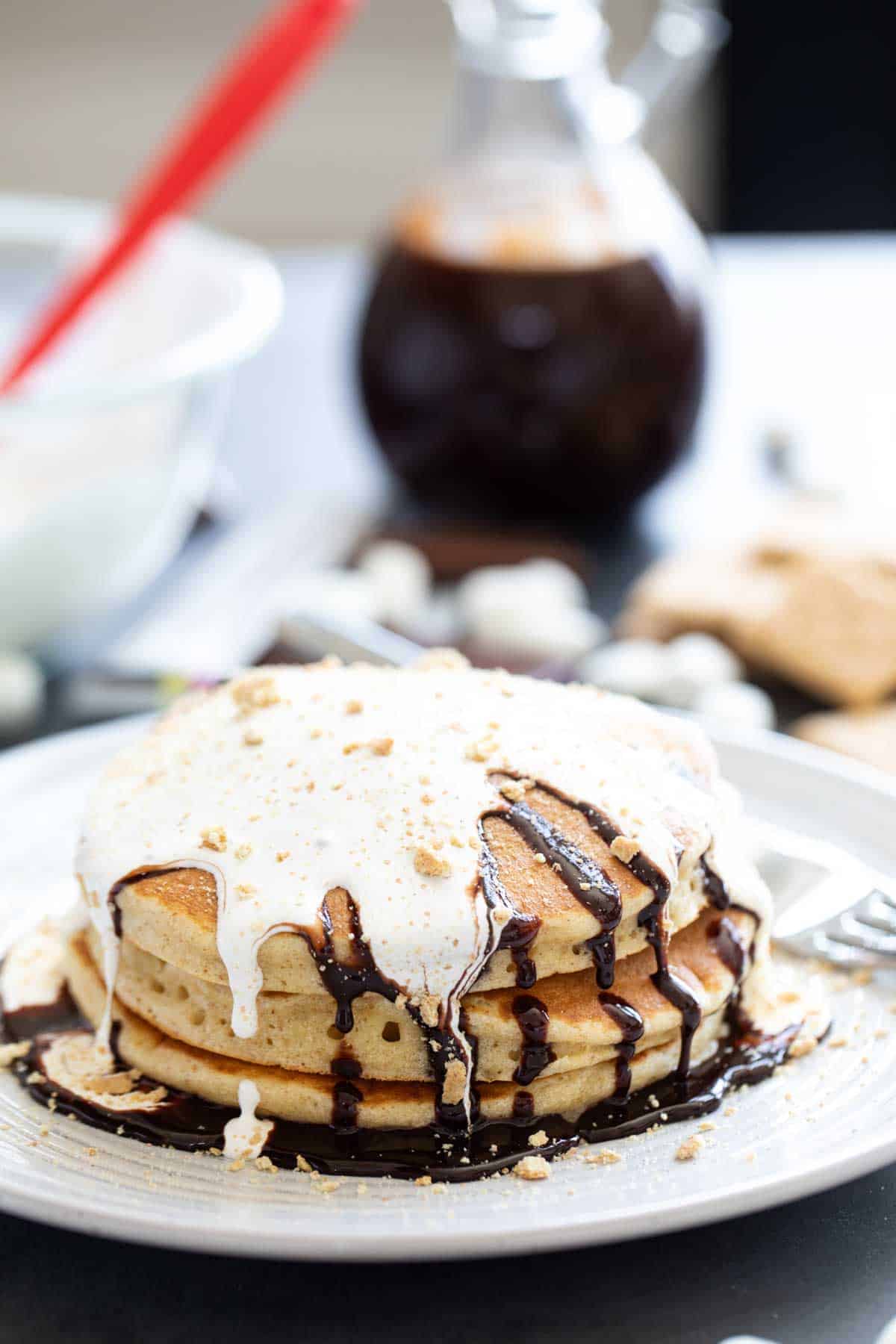 Stack of 3 s'mores pancakes topped with homemade chocolate sauce and marshmallow sauce with crushed graham crackers on top.