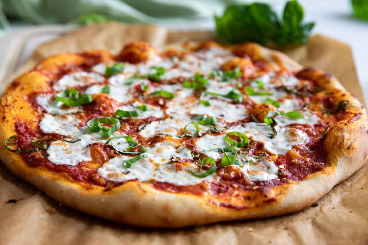 Full margherita pizza on a a piece of parchment paper with fresh basil in the background.